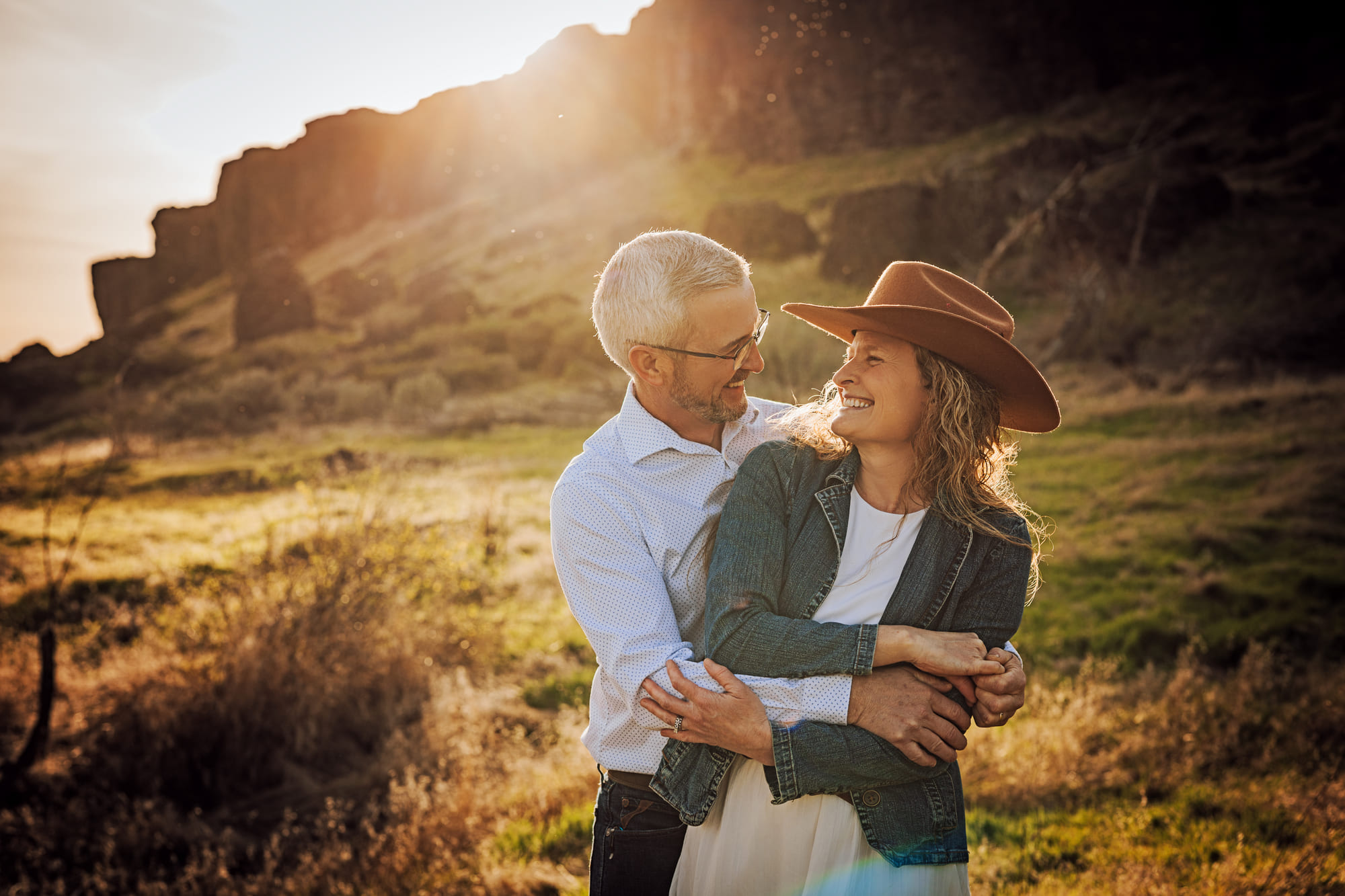 A couple laughing together in backlit evening light beside a rocky bluff.