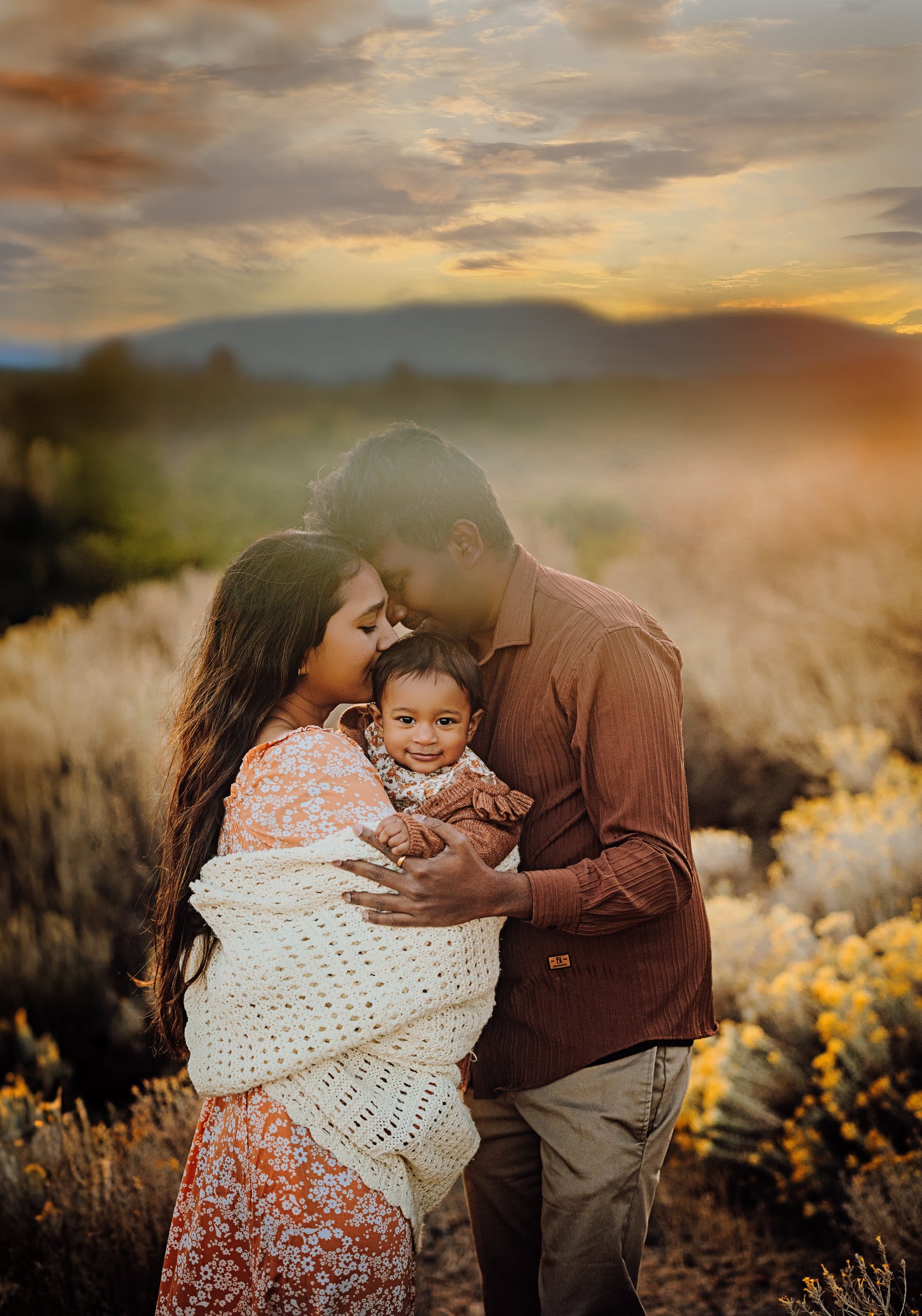 Two adults embracing their baby beneath a glowing evening sky.
