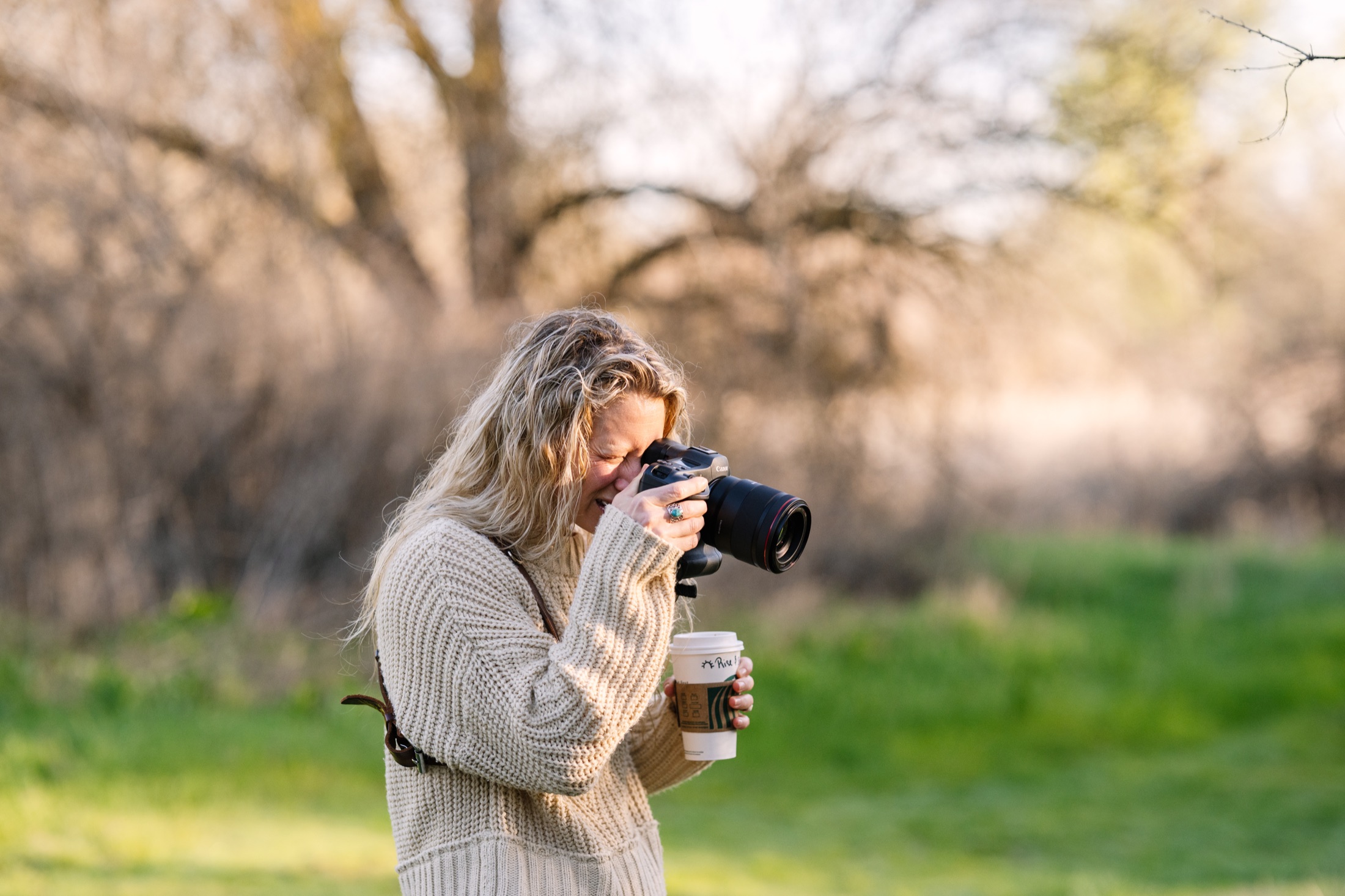 Lisa holding a camera to her eye and a takeaway cup while photographing outdoors.