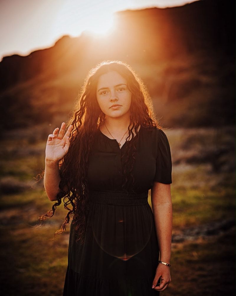 A senior in a black dress standing in dramatic golden-hour light