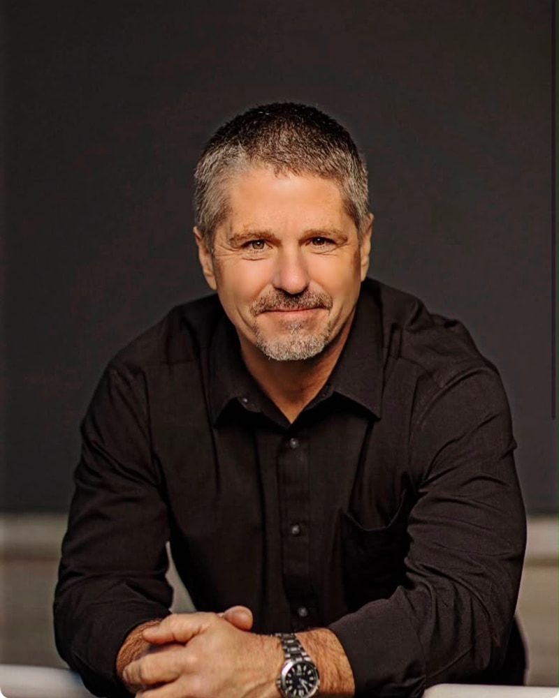 A man in a black shirt seated for a professional studio headshot